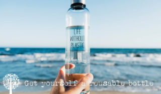 Una botella de agua reutilizable se sostiene frente a la cámara mientras las olas chocan contra la playa en el fondo.